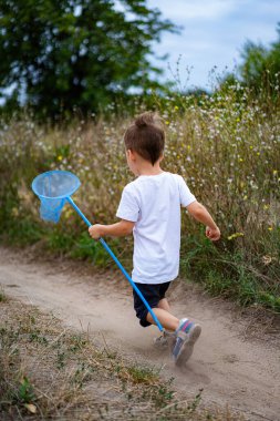 Çocuk yaz tarlasında ağ ile koşuyor. Genç bir çocuk neşeyle, çiçeklerle dolu güneşli bir yaz tarlasında kelebek ağı tutarak toprak bir patika boyunca koşar..