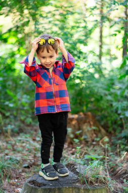 Gölgeli çocuk ormanda oynuyor. Neşeli bir çocuk ormanda bir kütükte durur, güneş gözlüğü takar ve doğada oynarken parlak bir ekose gömlek giyer..