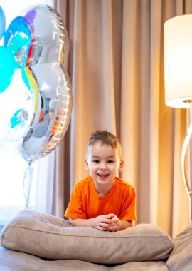 İçinde balonlar olan neşeli bir çocuk. Turuncu tişörtlü neşeli bir çocuk, yumuşak yastıklara yaslanırken gülümser. Etrafı renkli balonlar ve ılık ışıklarla çevrilidir..