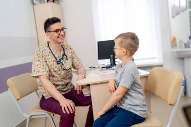 Genç hastası olan nazik bir doktor. Renkli önlüklü bir doktor, sağlık muayenesi sırasında gülümseyen bir çocuğu parlak bir muayenehaneye alır..