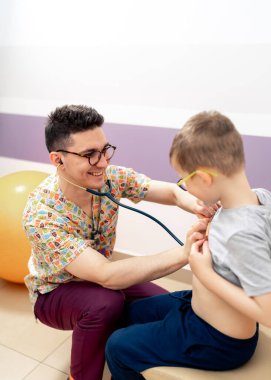 Doktor genç hastaya gülümsüyor. Bir doktor, tıbbi bir ofiste, dostane ve şefkatli bir çevreyi destekleyen genç bir çocuğu neşeli bir şekilde kontrol eder..