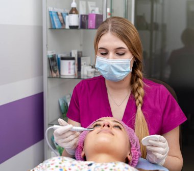 Cilt bakımında yüz bakımı. Bir cilt bakımı uzmanı, bir hastaya yüz tedavisi uygularken klinikte rahatlık ve güvenlik sağlar..