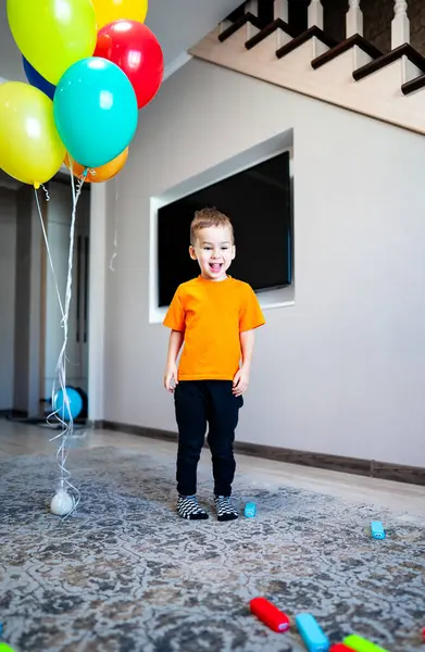 Neşeli çocuk balonlarla kutlama yapıyor. Turuncu tişörtlü enerjik genç, rahat bir oturma odasında renkli balonlarla neşeli bir anın tadını çıkarıyor..