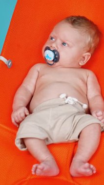 Sevimli beyaz bebek taklit edilmiş plajda dinleniyor. Şortlu, emzik takmış, dikkatlice bakan sevimli bir çocuk. Dikey video