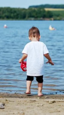 Beyaz tişörtlü sevimli çocuk nehre geliyor. Bebek su bidonunu suyla dolduruyor. Dikey video