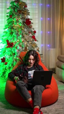 Kareli gömlek ve kot giyen bir kadın fasulye çuvalında oturuyor. Mutlu kız dizüstü bilgisayarda çalışıyor ve siyah kedisini tutuyor. Noel ağacı arka planda. Dikey video.