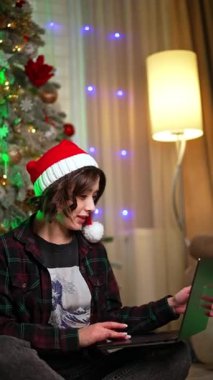 Noel Baba şapkası takan ve kareli gömlek giyen bir kız dizüstü bilgisayar tutuyor. Noel için internette çalışıyorum. Dikey video.