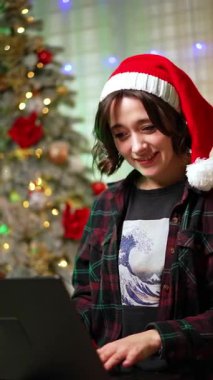 Kırmızı Noel Baba şapkalı gülümseyen kız dizüstü bilgisayarına odaklanmış. Tatillerde evden uzakta çalış. Noel ağacı arka planda. Dikey video.