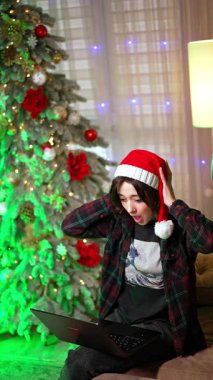 Şok olmuş Noel Baba şapkalı kız kafasını hayretle tutuyor. Evden çevrimiçi çalışmada başarı. Noel ağacı arka planda. Dikey video.