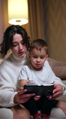 Beyaz kazaklı kadın beyaz tişörtlü şirin bir erkek çocuğu tutuyor. Anne ve oğlu akıllı telefondan konuşuyor. Dikey video.