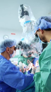 Baş cerrah bir ameliyat yapıyor ve mikroskobu hastanın üstüne çıkarıyor. Asistanı şırıngadan ilaç püskürtüyor. Dikey video.