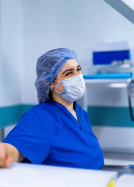 Mavi önlüklü, gülümseyen bir sağlık görevlisi. Maske takan ve mavi önlük giyen bir sağlık görevlisi steril bir tıbbi ortamda dururken gülümsüyor..