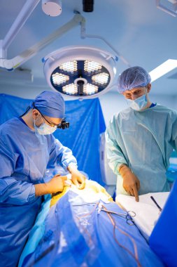 Cerrahi ekip operasyon halinde. Bir cerrahi ekibi ameliyat sırasında hastane ortamında parlak ışık altındaki bir hastayı ameliyat etmeye odaklanır..