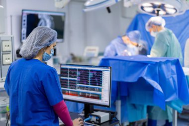 Cerrahi ekip iş başında. Bir tıbbi uzman bilgisayardaki hayati belirtileri gözlemlerken bir cerrahi ekip arka plandaki bir hastayı ameliyat ediyor..