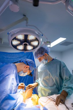 Cerrahlar hastanede iş başında. İki cerrah, bir ameliyathanede parlak cerrahi ışıkların altında odaklanmış ve hassas bir prosedürle meşgul..