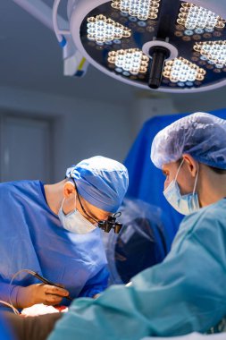Cerrahlar ameliyathanede. Cerrahlar, modern bir ameliyathanede hasta bakımına odaklanmış parlak ışıklar altında cerrahi müdahalede bulunurlar..