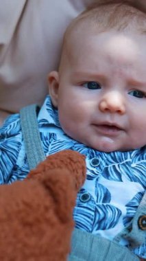 Mavi tişörtlü sevimli beyaz bebek yumuşak arka planda yatıyor. Yumuşak kahverengi ayıcık çocuğun yanında. Kapatın. Dikey video