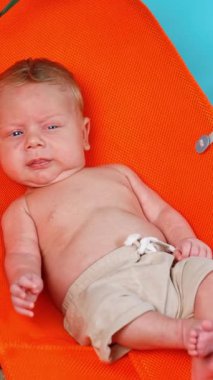 Mavi gözlü sarışın bir bebek sandalyede oturur. Doğaçlama kumsalda şortlu bir bebek dinleniyor. Dikey video