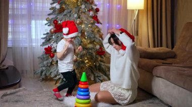 Kırmızı Noel Baba şapkası takan beyaz anne ve oğul Noel ağacının yanında yerde oturuyorlar. Bebek piramitle oynuyor ve annesi kepini düzeltiyor..