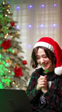 Laptopuna bakan neşeli esmer kız. Noel Baba şapkalı kadın heyecanla ellerini çırpıyor. Noel ağacı arka planda. Dikey video.