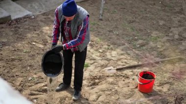 Kıdemli adam kovadan biraz su döküyor. Çiftçi bahçeye dikmek için genç bir ağaç alır..