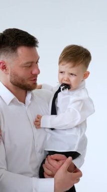 Baba oğul beyaz gömlek giyiyor. Küçük bebek ağlamaya başladı. Ebeveynler çocuklarını sakinleştiriyor. Dikey video.