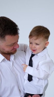 Beyaz gömlek giyen sakallı bir adam kucağında bir oğlan çocuğu tutuyor. Küçük çocuk babasının ellerinde inliyormuş. Dikey video.