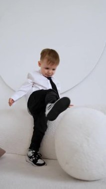 Beyaz gömlekli, siyah kravatlı, kot pantolonlu ve spor ayakkabılı sevimli beyaz çocuk kanepenin yanından geçiyor. Çocuk kanepedeki yastığı tekmeliyor. Dikey video.