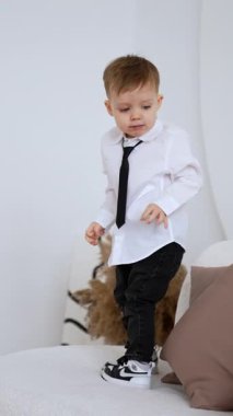 Beyaz gömlek giyen ve kanepede siyah kravat takan bir çocuk yürüyor. Küçük çocuk yumuşak pamuk yumağıyla oynuyor. Dikey video.