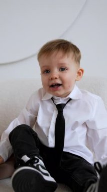 Sevimli, gülümseyen kızıl saçlı, kravat takan erkek bebek kanepede oturuyor. Gülümseyen çocuk kanepede oturur ve ayağa kalkıp koşar. Dikey video.