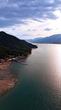 Güneşte parlayan yırtık pırtık yüzeyi olan muhteşem bir su manzarası. Sahilde ormanlarla kaplı muhteşem dağlar var. Gökyüzünü kaplayan dramatik bulutlar. Hava görüntüsü. Dikey video.