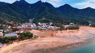 Japonya 'nın Miyajima adasındaki İç Denizi' nin kumlu kıyısı. İnsanlar Itsukushima Tapınağı 'ndaki ikonik Grand Torii Kapısı' nın yanında yürüyorlar. Yeşil dağların eteklerindeki çoklu evlere bakın..