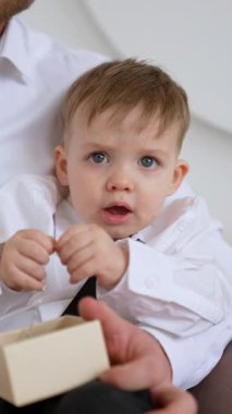 Beyaz tişörtlü beyaz erkek bebek babasının elinde oturuyor. Küçük çocuğun elinde gümüş bir zincir var. Dikey video.