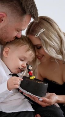 Siyah pasta yiyen sevimli beyaz bir bebek. Sevgi dolu ebeveynler ve oğulları çocuklarının doğum gününü kutluyorlar. Dikey video.