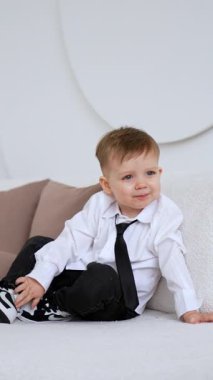 Beyaz gömlekli, siyah kotlu ve spor ayakkabılı beyaz erkek bebek kanepede oturuyor. Mutlu çocuk ayağa kalkar ve yastığı atar. Dikey video.