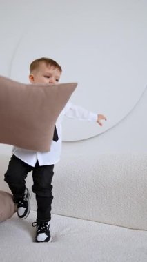 Kanepede gömlek ve kravat takan beyaz bir çocuk duruyor. Mutlu çocuk minderlerle oynuyor. Dikey video.