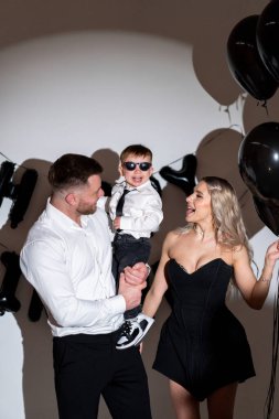 Balonlar ve eğlence ile aile kutlaması. Siyah balonlarla çevrili güneş gözlüklü bir çocuğu tutan bir erkek ve kadınla neşeli bir aile anı..