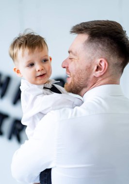 Baba ve oğul neşeyi paylaşır. Genç oğlunu kucaklarken gülümseyen bir adam, beyaz gömlek ve papyon takıyor, neşeli bir atmosferde..