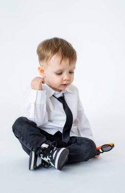 Yerde kravatlı bir çocuk var. Oyuncu çocuk yerde oturuyor, gömlek ve kravat takmış, merakla güneş gözlüğünü inceliyor..