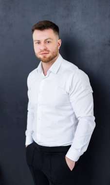 Profesyonel bir adam beyaz gömlekle poz verir. Beyaz gömlekli bir adam, elleri cebinde karanlık bir duvara yaslanmış, kendinden emin bir şekilde duruyor..