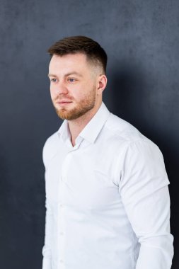 Hafif gömlekli sakallı adam. Kısa sakallı bir adam koyu bir arka plana karşı duruyor, şık bir elbise içinde düşünceli bir şekilde yan tarafa bakıyor..