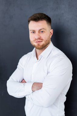 Kendine güvenen adam içeride poz verir. Bakımlı sakallı, kendine güvenen bir adam kollarını beyaz bir gömlekle koyu bir fonda kavuşturur..