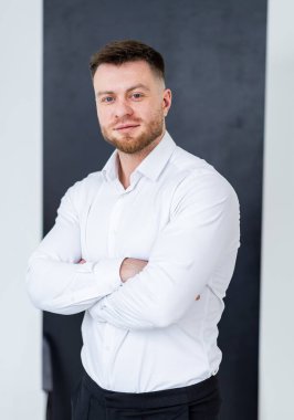 Modern bir ofisteki adam. Kısa saçlı ve sakallı bir adam şık bir ofiste duruyor, beyaz gömlek ve siyah pantolon giyiyor..