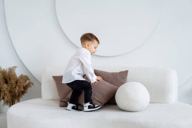 Çocuk evin içindeki kanepeye tırmanıyor. Beyaz tişörtlü bir çocuk şık bir kanepede oynuyor. Parlak, minimumda, dairesel dekorlu..