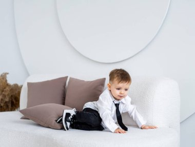 Takım elbiseli çocuk kanepede oynuyor. Resmi tişört giyen ve kravat takan genç bir çocuk, parlak bir odada şık bir kanepede minderlerle şakalaşarak etkileşime geçer..