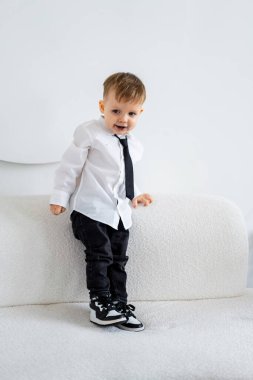 Takım elbiseli çocuk fotoğraf için poz veriyor. Beyaz gömlekli ve siyah kravatlı neşeli bir genç bir çocuk kanepede oturur ve oyuncu kişiliğini gösterir..