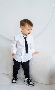 Güzel giyimli genç çocuk. Beyaz gömlekli ve siyah kravatlı genç bir çocuk zarif bir şekilde beyaz bir kanepenin yanında duruyor, şık bir elbise sergiliyor..