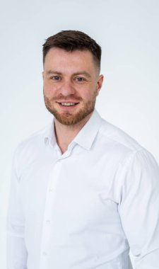 Beyaz gömlekli profesyonel adam gülümsüyor. Düzgün sakallı ve kısa saçlı bir adam sade bir arka planda duruyor, beyaz gömlekle güvenle gülümsüyor..