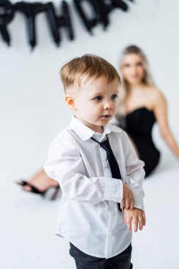 Doğum günü partisinde takım elbiseli bir çocuk. Beyaz gömlekli ve siyah kravatlı genç bir çocuk bir doğum günü partisinde duruyor ve arkasında bir kadın oturuyor..
