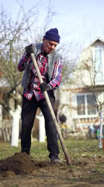 Yaşlı beyaz erkek, kareli gömlek ve yelek giyiyor. Evin yakınındaki zemini kazıyor. Çiftçi bir delik açar ve küreği toprağa saplar. Dikey görünüm.